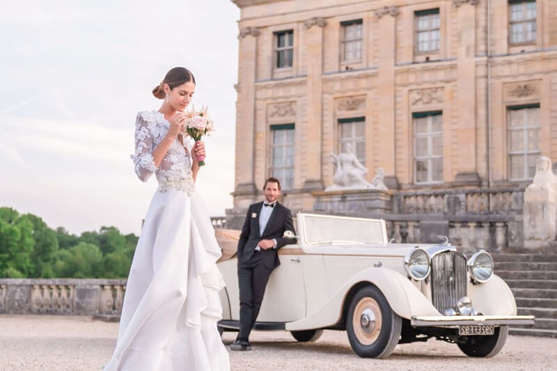 Luxurious French chateau wedding at Vaux le Vicomte, photographed by elite destination wedding photographer, Kiss Me in New York, and showcasing a stylish couple posing against the backdrop of a vintage car and the castle.