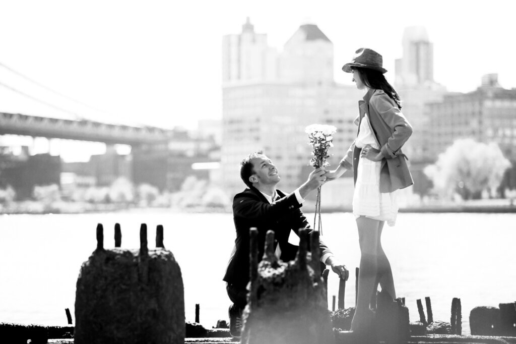 Love-filled engagement photograph using a picturesque setting under the Brooklyn Bridge, NYC, with a gentleman offering his fiancée a bouquet of fabulous flowers.