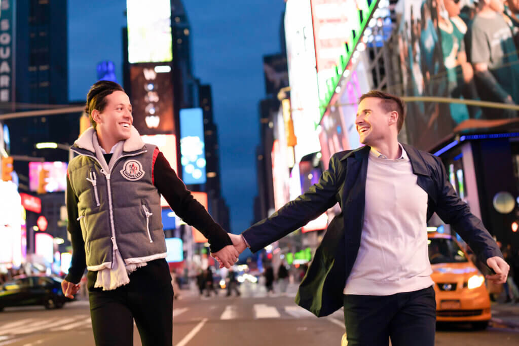 Same-sex couple running in the middle of Times Square at night, playfully embracing the bright lights and energy of New York City.