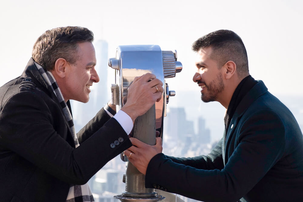 Same-sex couple engagement photo session on the Top of The Rock observation deck, overlooking panoramic views of the New York City skyline as the backdrop.