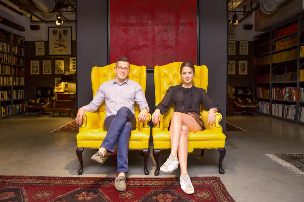 Fashionable couple sitting side by side on two vibrant yellow chairs during an editorial style NYC Engagement photo session.