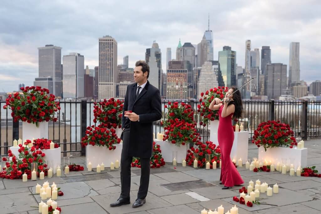 A stylish gentleman stands poised against the spectacular NYC skyline, moments before dropping to one knee to set the scene for a fairytale winter proposal. Romance fills the air as countless red rose petals encircle the setting, and the melodic serenade of a master violinist adds an enchanting touch.